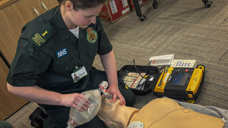 Woman wearing green ambulance uniform with apprentice medical technician written on her epaulette, is placing a resuscitation mask over a mannequin.
