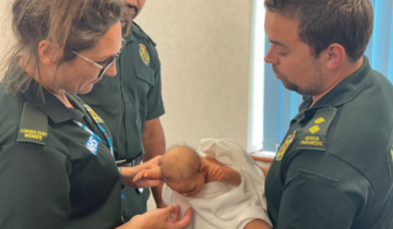 Two clinicians in ambulance uniform gently hold a newborn wrapped in a white blanket. The uniforms display NHS logos, and badges indicate roles such as Senior Paramedic and Consultant Midwife. They're standing in a room with natural light coming through blinds.