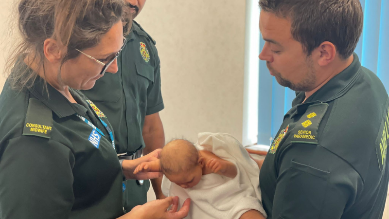 Two clinicians in ambulance uniform gently hold a newborn wrapped in a white blanket. The uniforms display NHS logos, and badges indicate roles such as Senior Paramedic and Consultant Midwife. They're standing in a room with natural light coming through blinds.