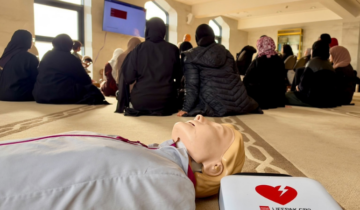 CPR training at a Mosque with people sat on a wooden floor back facing the camera and a resuscitation training manikin and defibrillator visible on the floor.