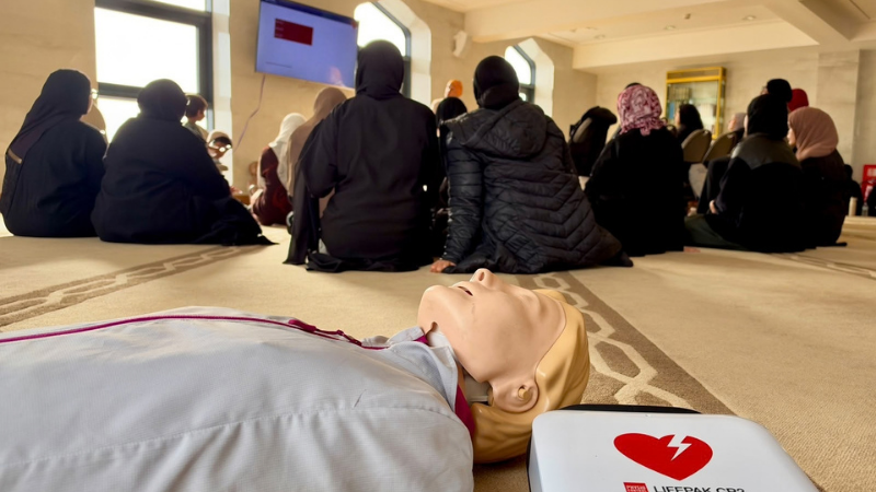 CPR training at a Mosque with people sat on a wooden floor back facing the camera and a resuscitation training manikin and defibrillator visible on the floor.
