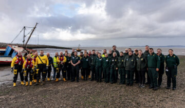 Large group people wearing green ambulance uniform alongside other wearing Lifeboat response equipment. They standing on a mudbank with a beached boat behind them.