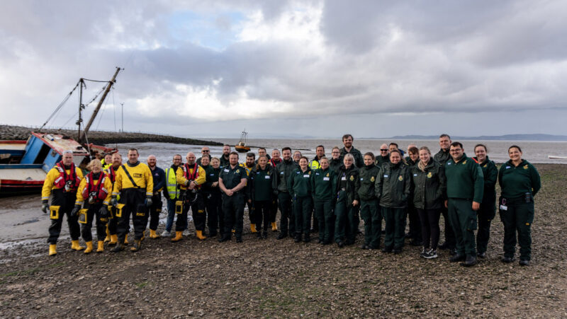 Large group people wearing green ambulance uniform alongside other wearing Lifeboat response equipment. They standing on a mudbank with a beached boat behind them.