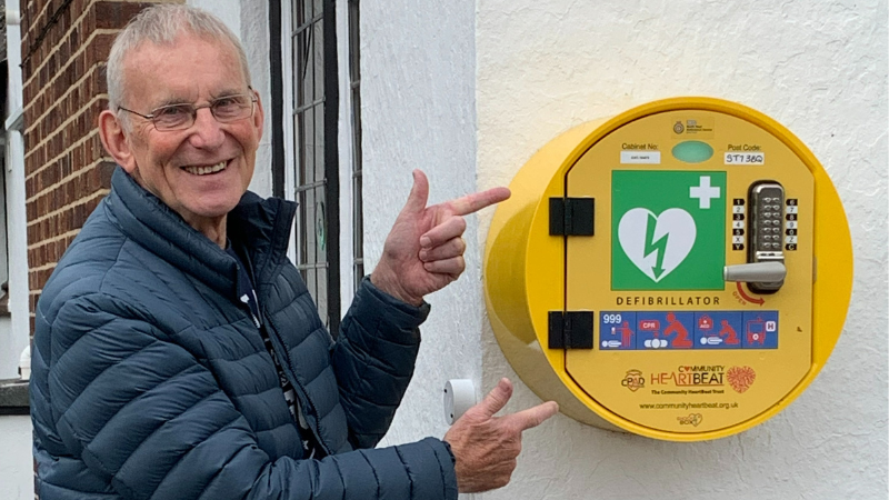 Volunteer Joe pointing to a wall mounted defibrillator