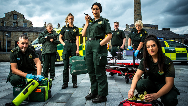 Male and female ambulance crew mates and colleagues in uniform stood on a pavement with kit bags looking at the camera.