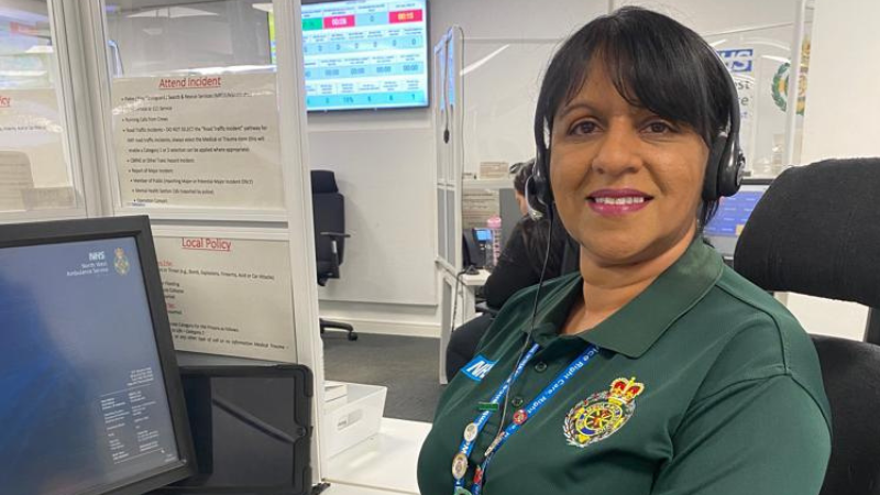 Emergency Medical Advisor Heena who is female and is wearing a headset in her uniform. She is sat down smiling at the camera. There is a computer on the left of the image.