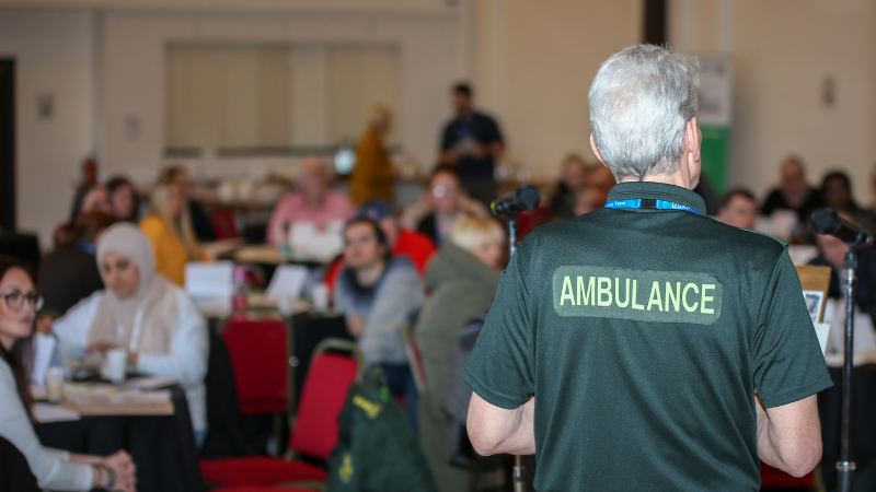 A man wearing green, talking to audience, sat around tables. The man has the word ambulance written on the back.